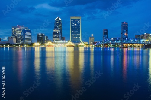 View of a downtown at dusk with buildings, bridge and colored lights reflecting over water shot as long exposure from across the water with blue sky, landscape composition