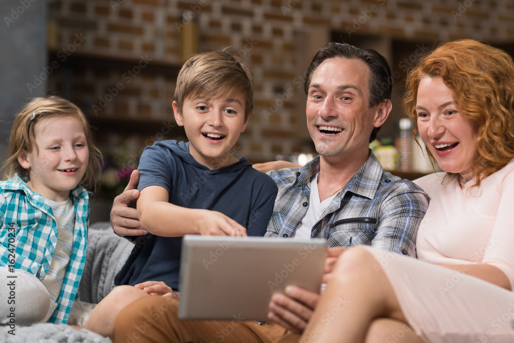 Happy Smiling Family Use Tablet Computer Sitting On Couch In Living Room, Parents Spending Time With Son And Daughter Surfing Internet Happy Smiling Family Use Tablet Computer Sitting On Couch In Living Room, Parents Spending Time With Son And Daughter Surfing Internet