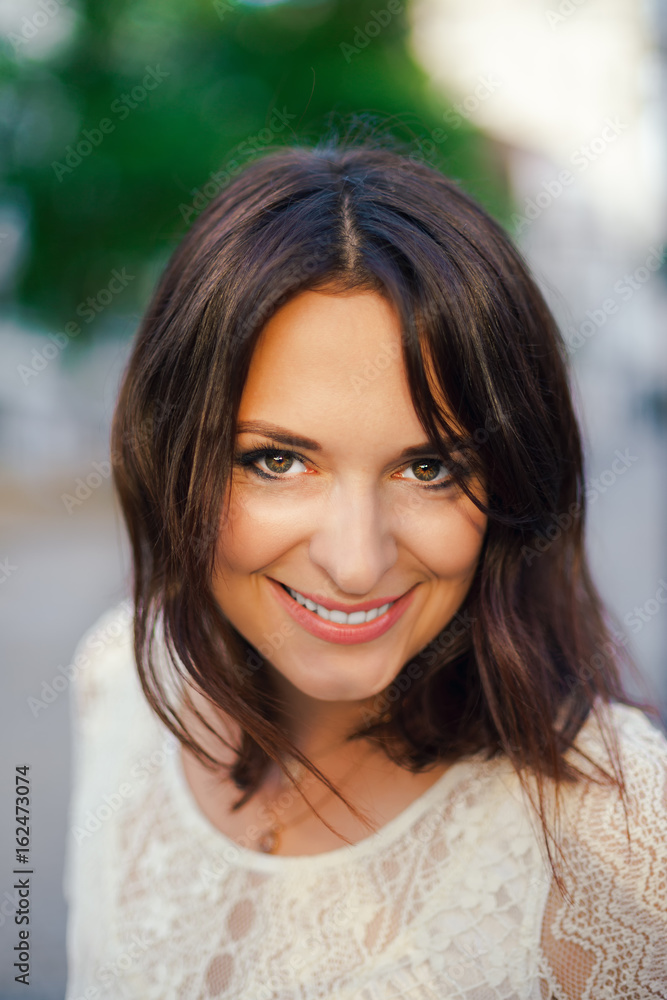 Young beautiful woman, dark-haired, smiling, peering into the frame, visiting the city center on a sunny day, in a white dress from the street in the background. Outdoor portrait. Young beautiful woman, dark-haired, smiling, peering into the frame, visiting the city center on a sunny day, in a white dress from the street in the background. Outdoor portrait.