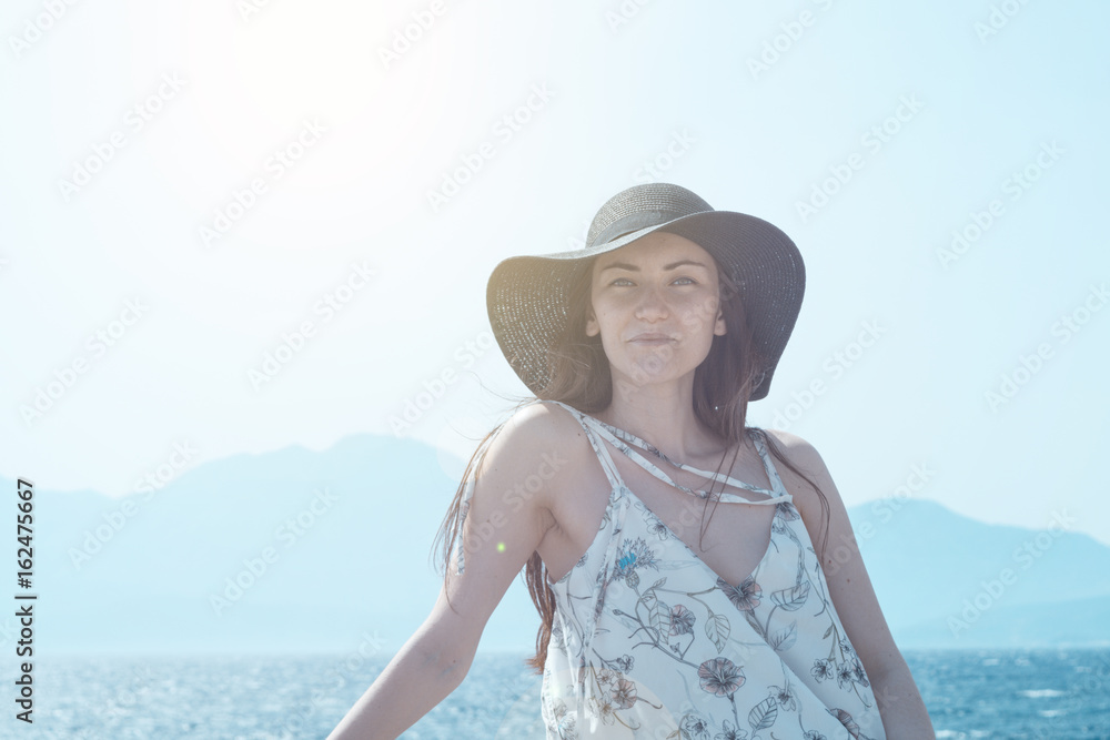 Outdoor summer portrait of young pretty woman looking to the camera in the port of Agios Nikolaos, enjoy her freedom and fresh air, wearing stylish hat and clothes