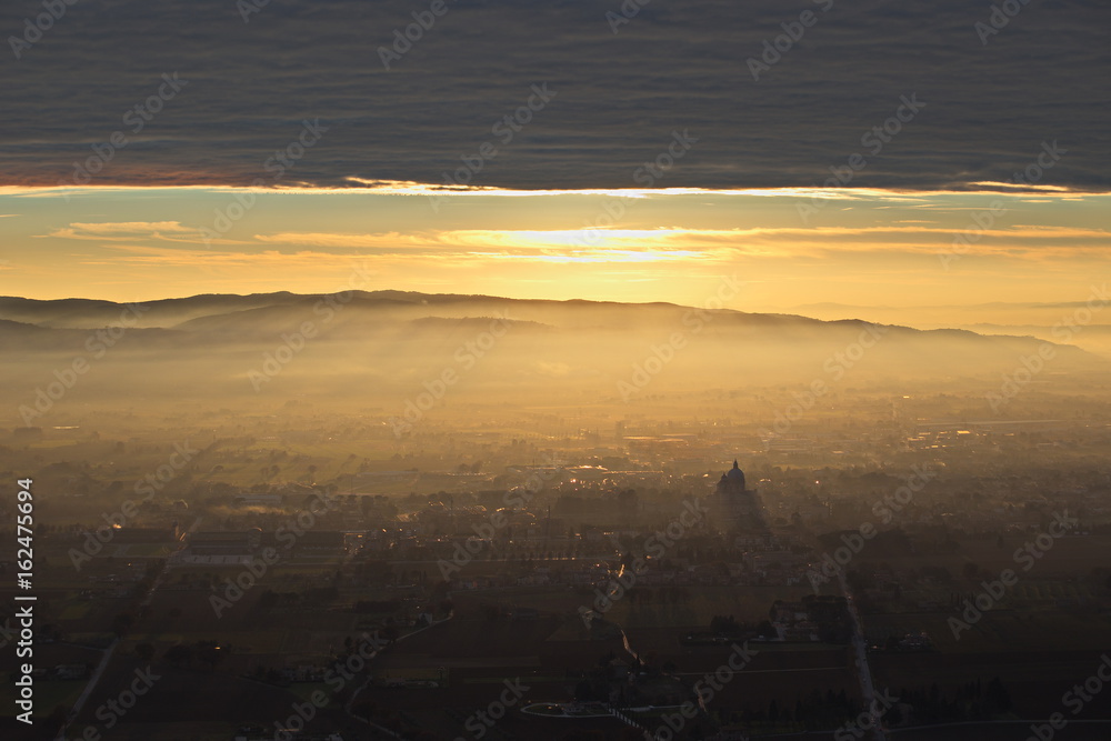 An epic, aerial view of Santa Maria degli Angeli town (Assisi) , with the sun coming down through a rooftop of clouds, projecting sunrays, and lighting up half of the valley