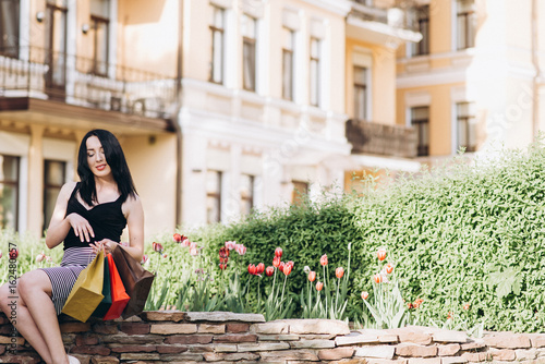 Fashionably dressed woman with colored shopping bags sitting near flowers on the streets and looking into the bag, shopping concept © Anastasiia