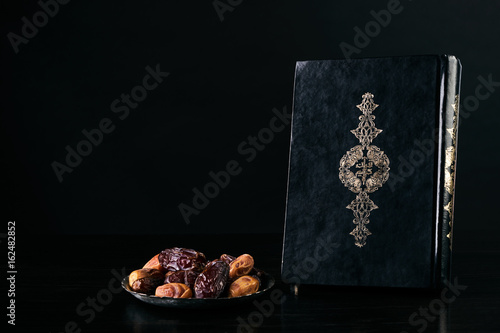 Ramadan atmosphere, Quran/ Koran Kareem book and silver dish with dry dates isolated on black background with copy space.
