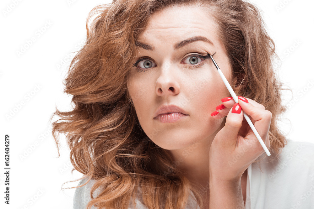 focused woman looking at camera while putting eyeliner isolated on white focused woman looking at camera while putting eyeliner isolated on white