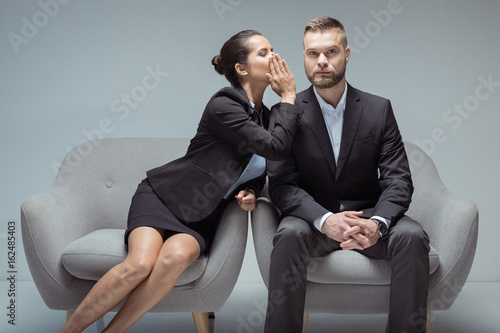 businesswoman whispering something on colleague's ear while sitting on chairs photo