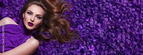The girl in the petals. Beautiful young girl lies in the violet petals in a long dress. Glamor, luxe. Hair - curls. Makeup - arrows, purple lipstick. Love, romance.