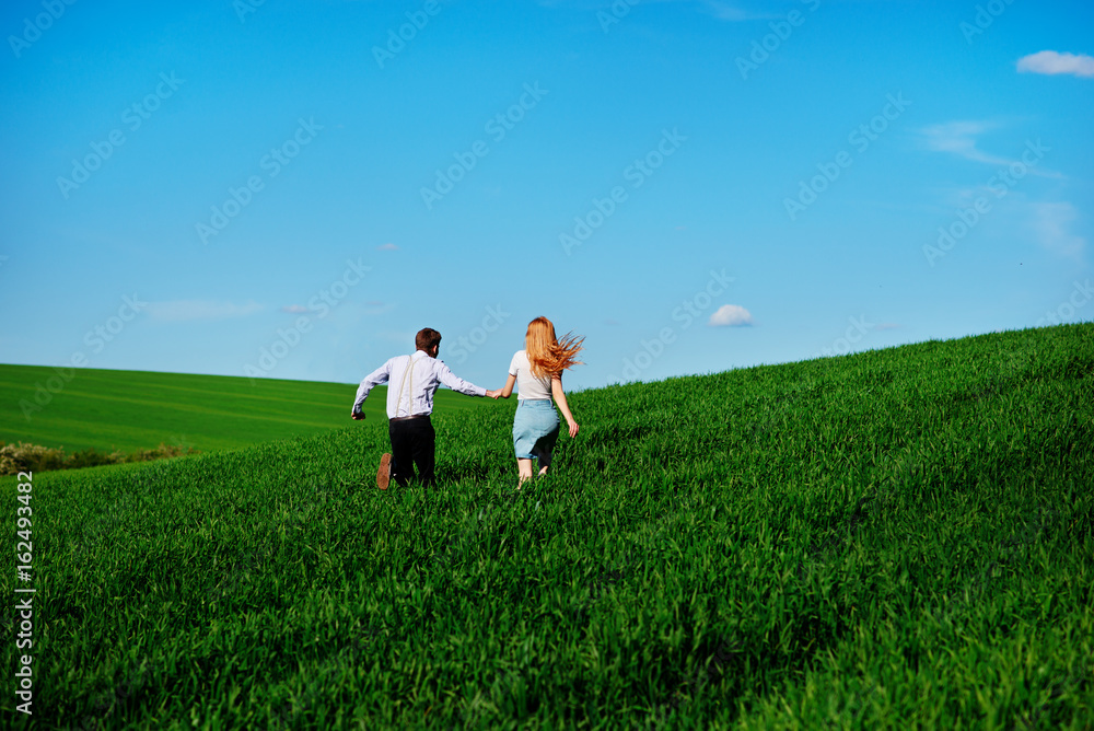 Young happy lovers running on meadow with green grass and blue sky, From the back Young happy lovers running on meadow with green grass and blue sky, From the back