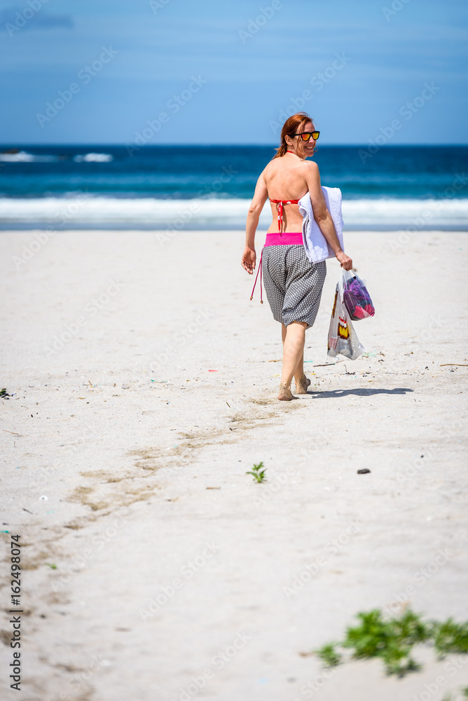 Woman walking the beach to ocean carrying towel and bags.