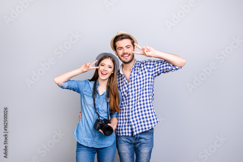Family memories. Vacation together. Happy young couple in casual wear and hats are embracing on light background, girl is holding digital camera, smiling, gesturing vsigns