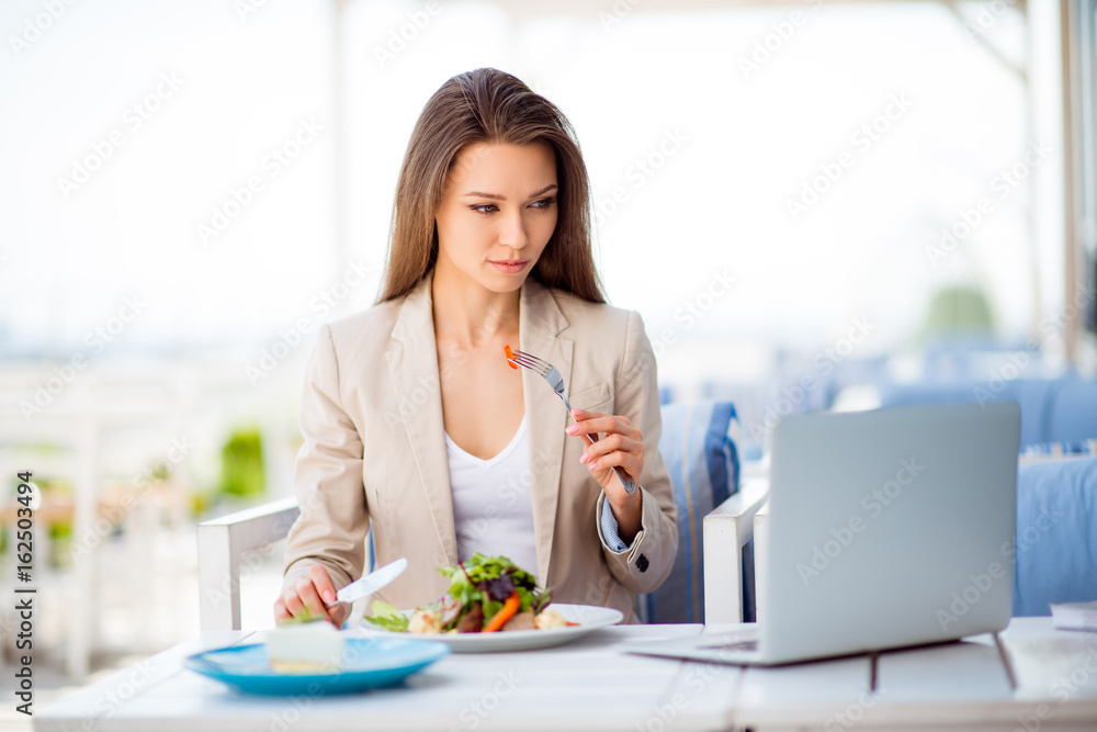 Pensive young business lady is having her lunch at the roof top restaurant`s open air light terrace, she is in a formal clothes, looking at the screen of her laptop