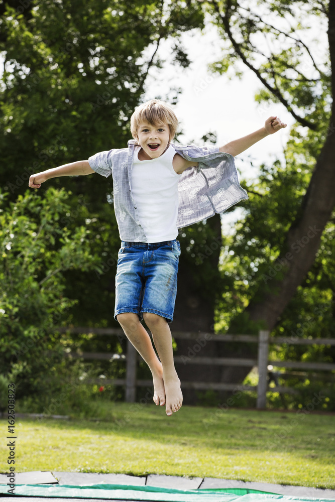 Boy wearing shirt and denim shorts jumping on a trampoline in a garden. Boy wearing shirt and denim shorts jumping on a trampoline in a garden.