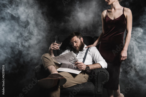 stylish man holding whisky glass and reading newspaper while woman standing near him © LIGHTFIELD STUDIOS