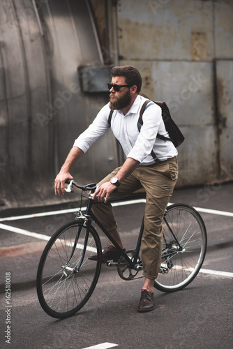 serious stylish bearded man in sunglasses riding bicycle