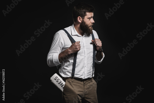 fashionable man in white shirt and suspenders standing with business newspaper in pocket isolated on black