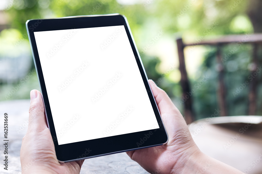 Mockup image of woman's hands holding black tablet pc with white blank screen on vintage wooden table and green nature background
