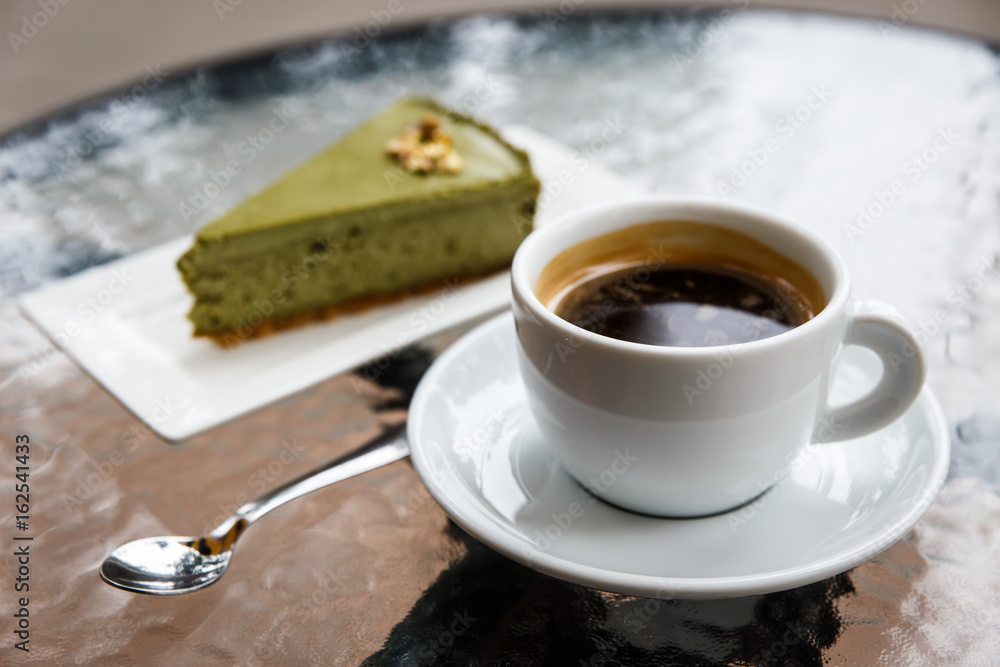 A pistachio cheesecake on a plate, a white cup of black coffee, a tea spoon. On a glass table top. Selective focus. A pistachio cheesecake on a plate, a white cup of black coffee, a tea spoon. On a glass table top. Selective focus.