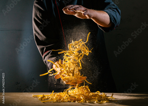 Chef adds wheat flour to the Italian ribbon-shaped pasta, before preparing the dish. Freeze motion effect. Strong contrast side lighting.