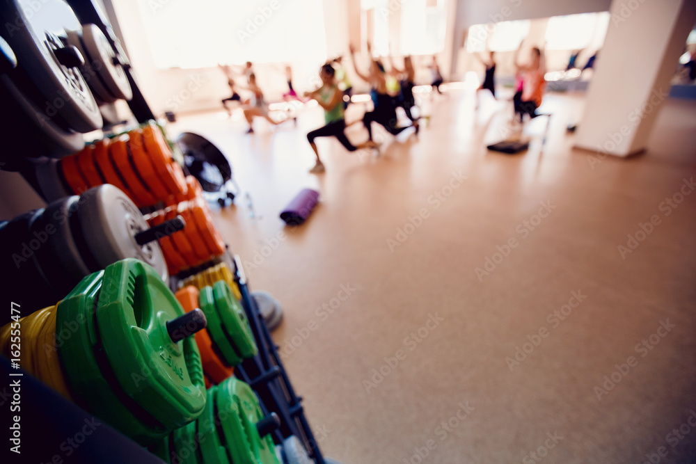 Obraz premium In the foreground a sports projectile disc for a bar, in the background a group of girls of friends are engaged in fitness. Sports hall concept gym. Glare sun