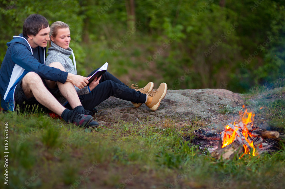 young couple sitting near campfire with book at camp in woods. C