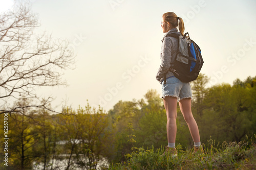 Hiker woman takes a rest during summers expedition. Girl is looking around. River bank and forest background. Travel, vacation, holidays and adventure concept. © sasapanchenko