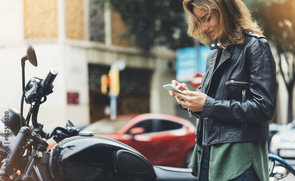 Girl in leather jacket holding smart phone on background motorcycle in sun city, hipster using in female hands and texting mobile, glitter street lifestyle, tourist planing route in summer concept