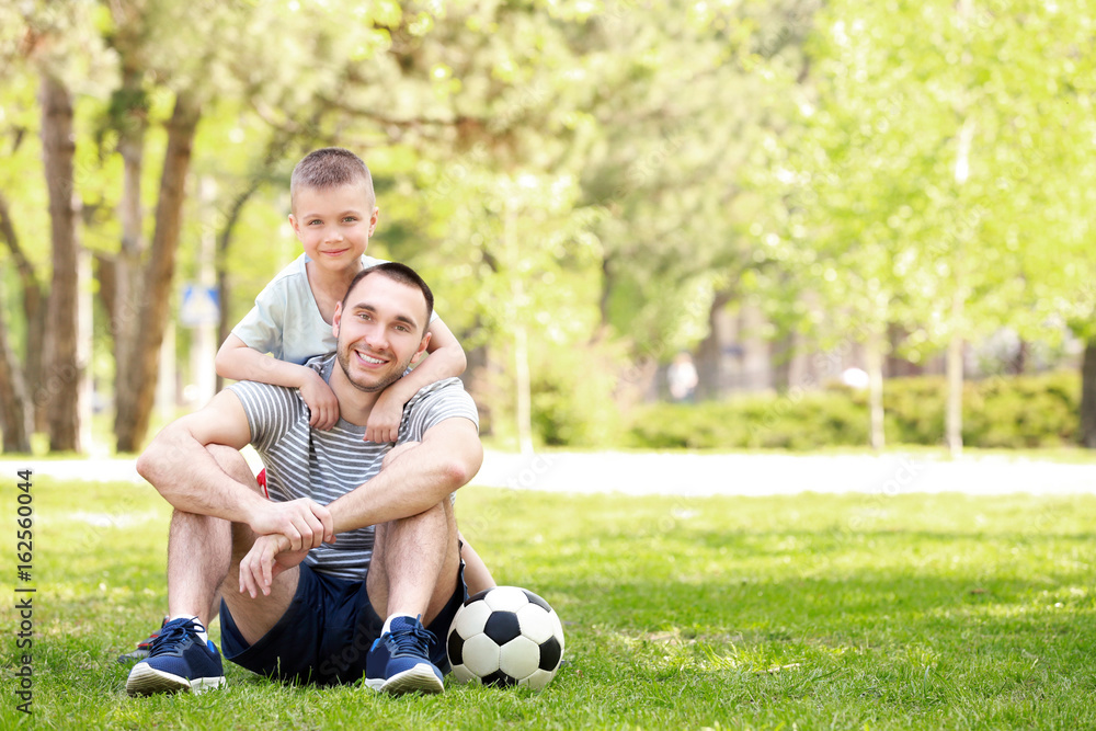 Father and son with soccer ball sitting on green grass in park