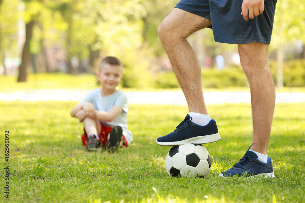 Young man with soccer ball in green park and his blurred son on background