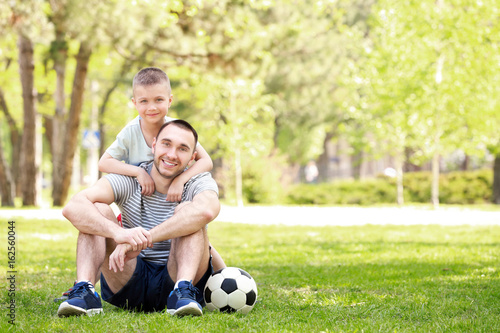 Father and son with soccer ball sitting on green grass in park © Africa Studio