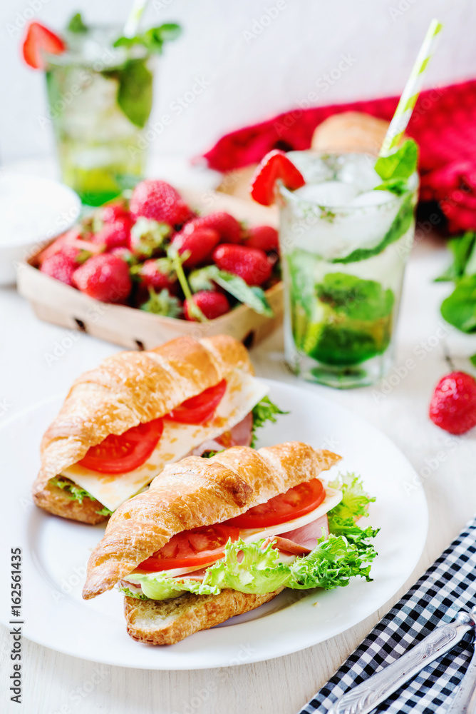 Breakfast, business lunch, sandwiches croissant with ham, cheese, sauce, lettuce, tomato and strawberry mint ice-cold cocktail on a light background Breakfast, business lunch, sandwiches croissant with ham, cheese, sauce, lettuce, tomato and strawberry mint ice-cold cocktail on a light background