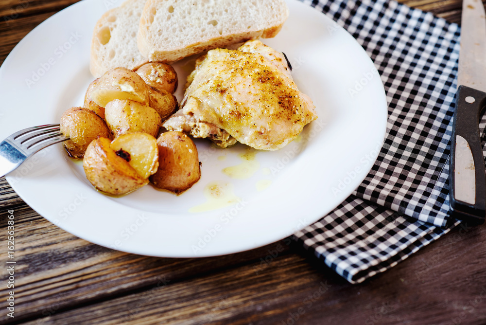 Hearty and delicious dinner, baked chicken legs and thighs with potatoes, onions, tomatoes, spices, herbs and white bread on a dark wooden background