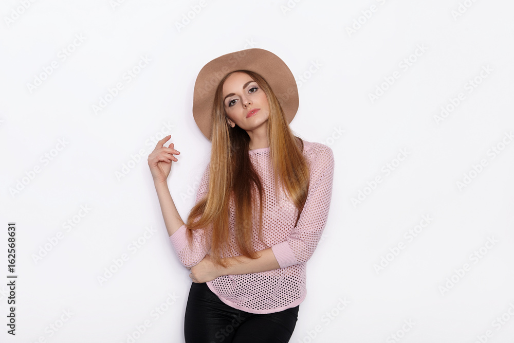 Cute and stylish beautiful young blonde woman in trendy clothing practicing model poses and looking away with smile while standing against white wall background Cute and stylish beautiful young blonde woman in trendy clothing practicing model poses and looking away with smile while standing against white wall background
