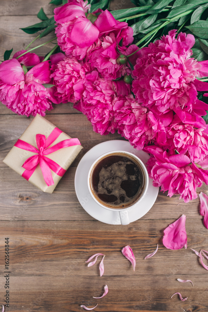 A cup of coffee , pink peonies pattern and gift box on wooden background . Good morning. Women's or Mother's Day background.