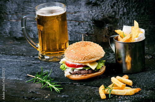 Hamburger with french fries, beer on a burnt, black wooden table. Fast food meal. Homemade hamburger consist of beef meat, lettuce, tomato, bins, dressing, cheese and spices. Vintage 
