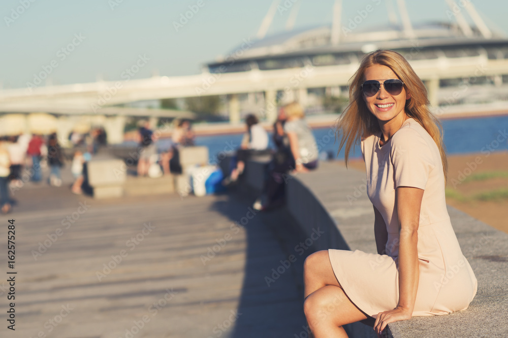 Pretty young blonde woman sitting at the seashore looking at camera and smiling
