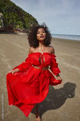 Female dark skin model with curly hair in red dress enjoing at the sea coast