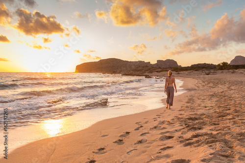 Woman walking on sandy beach at golden hour. Seashore sunset walk, Falasarna, Crete, Greece.