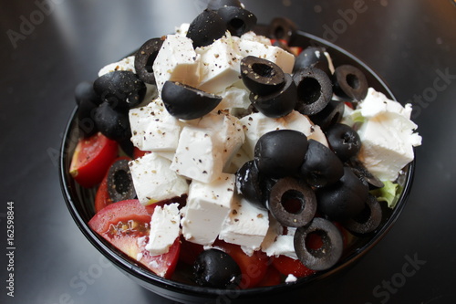Greek salad with sirtaki cheese and black olives and a red tomato. On white cheese is ground pepper. Salad in a glass plate is on a dark table.