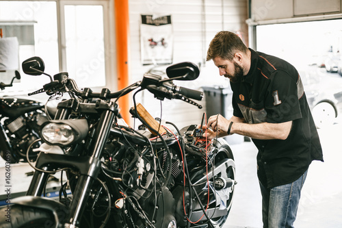 Professional motorcycle mechanic working in bike repair service. Mechanic checking a bike battery level with voltmeter in garage