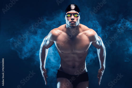Attractive and muscular swimmer. Studio shot of young shirtless sportsman on black smoke background. Man with glasses photo