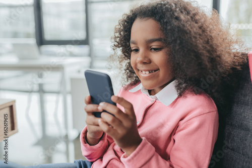 little happy african american girl with curly hair using smartphone