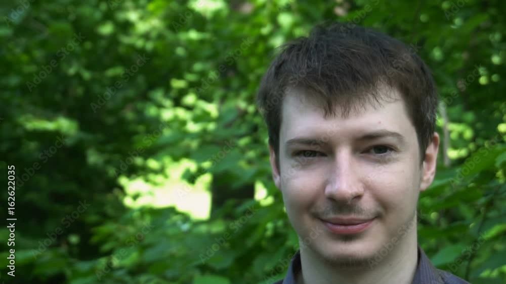 View on a young handsome man who looks satisfied, stare into camera and stands in the park . Slow motion. View on a young handsome man who looks satisfied, stare into camera and stands in the park . Slow motion.