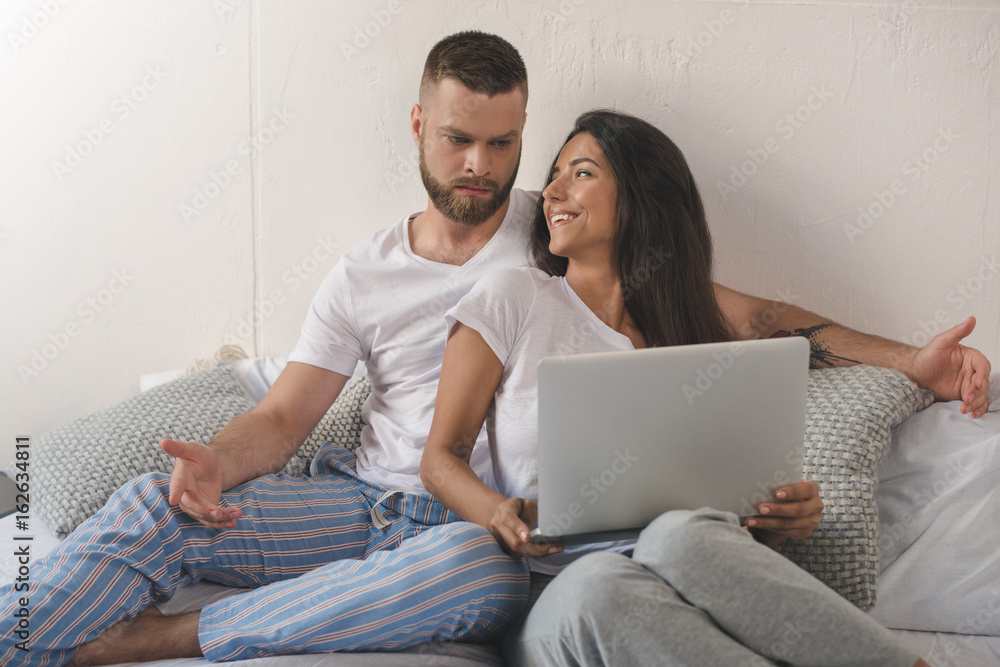 smiling young woman using laptop while her dissatisfied boyfriend gesturing in bed