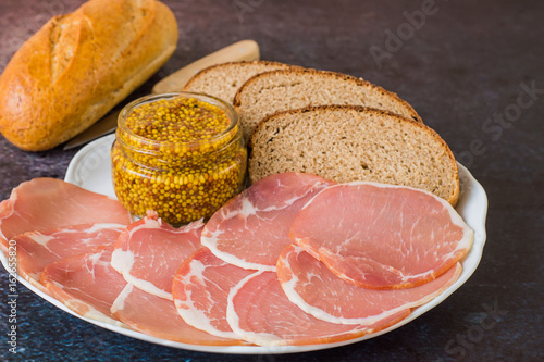 Fototapeta Naklejka Na Ścianę i Meble -   Fast snack. Smoked bacon, sliced thin slices, bread and mustard on a white porcelain dish on a dark background.