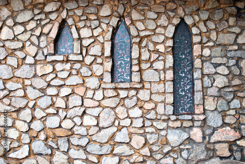 Stone gray wall of large stones with different shapes and textures with three narrow windows with patterns on the glass in the shade of trees on a sunny summer day