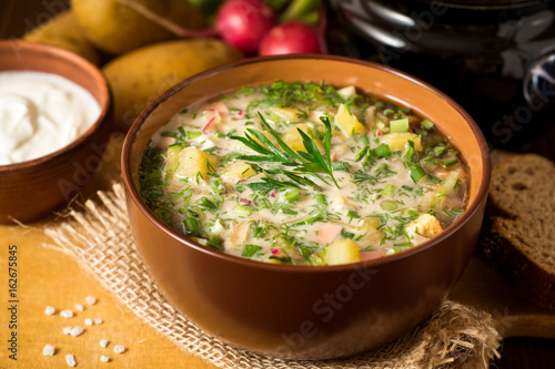 Okroshka. Traditional Russian summer cold soup with sausage, vegetables and kvass in bowl on wooden background.