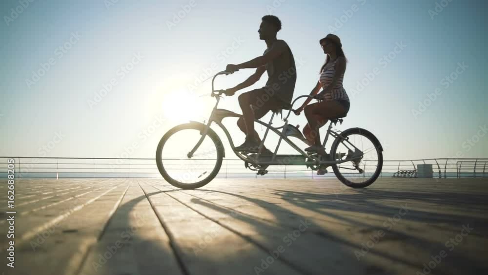 Portrait of a mixed race couple riding on tandem bicycle outdoors near the sea, slow motion Portrait of a mixed race couple riding on tandem bicycle outdoors near the sea, slow motion