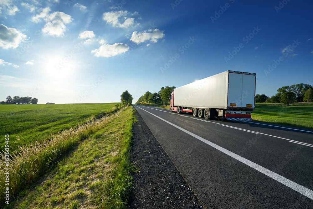 Red truck driving on asphalt road along the green fields in rural landscape at sunset