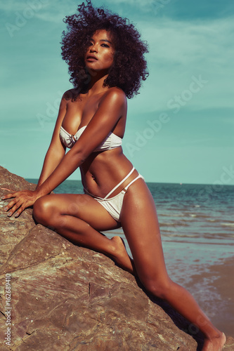 Exotic model with tanned skin and curle hair in bikini posing at the rock beach