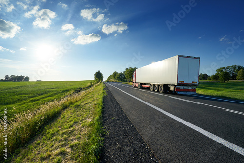 Obraz na płótnie Red truck driving on asphalt road along the green fields in rural landscape at sunset w salonie