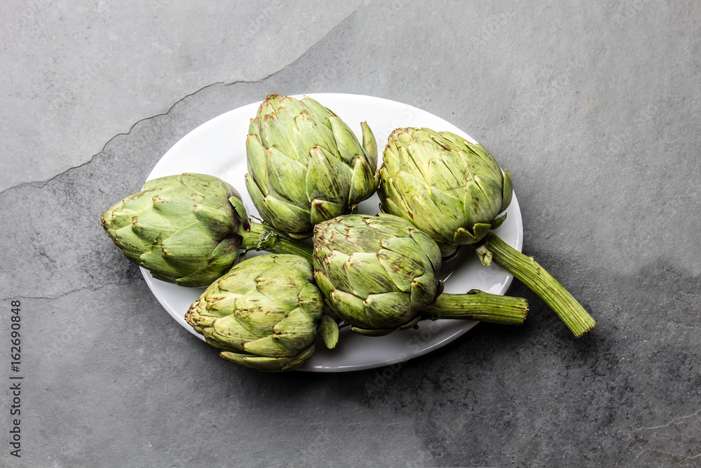 Fototapeta premium Fresh artichokes on withe plate, slate background